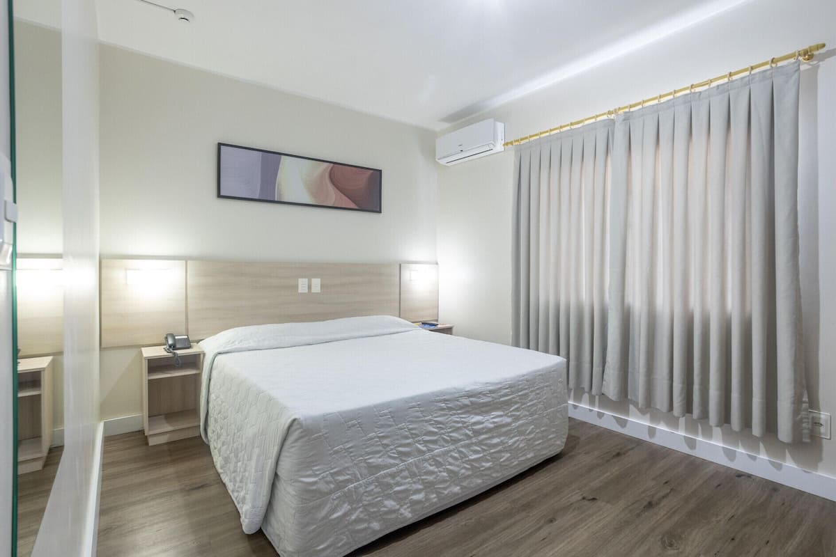 A hotel room with a bed, bedside tables, an air conditioning unit, a painting on the wall, and a window with curtains.