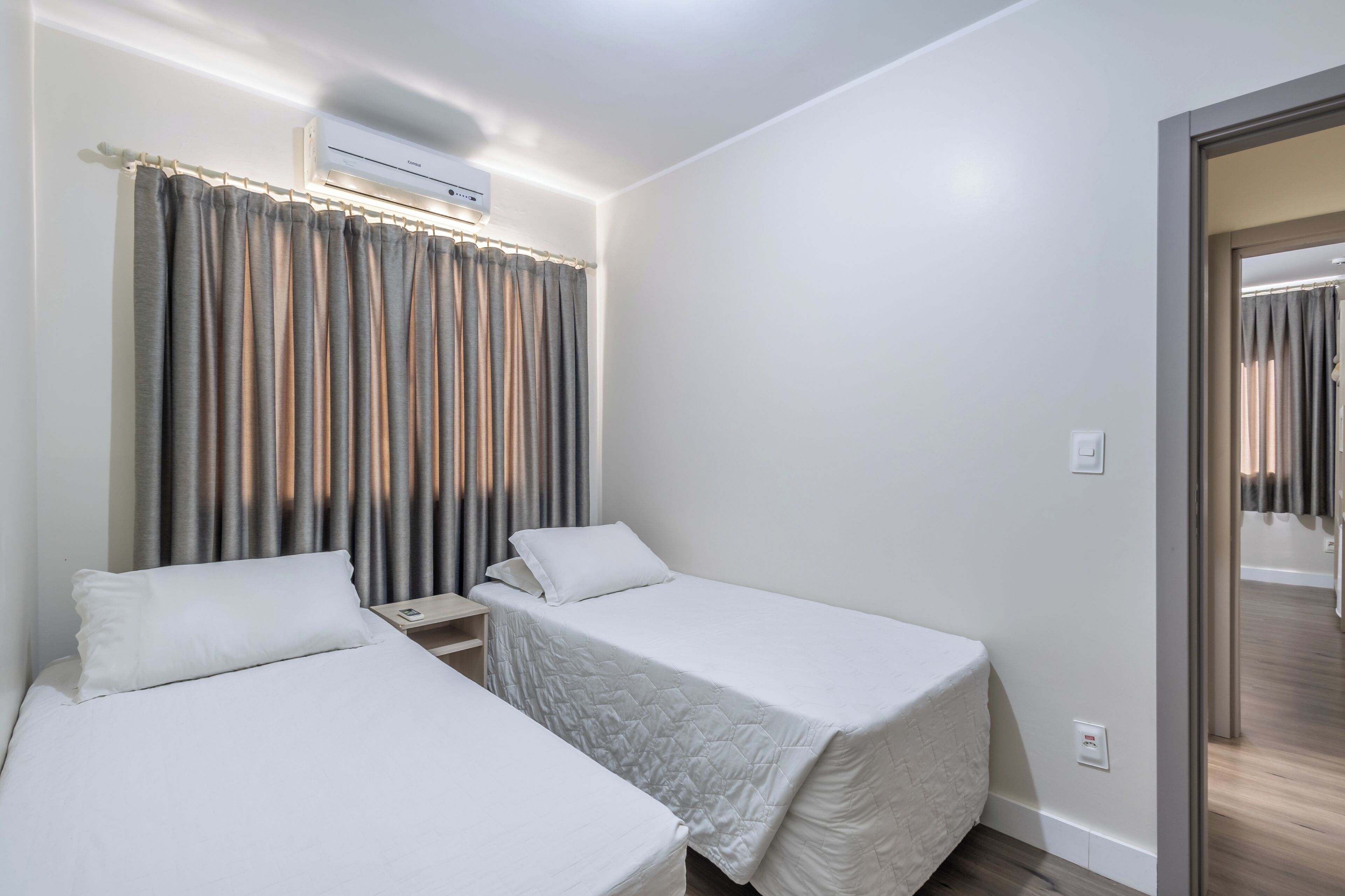Habitación de hotel con dos camas individuales, una mesita de noche, aire acondicionado instalado en la pared y una ventana con cortinas.