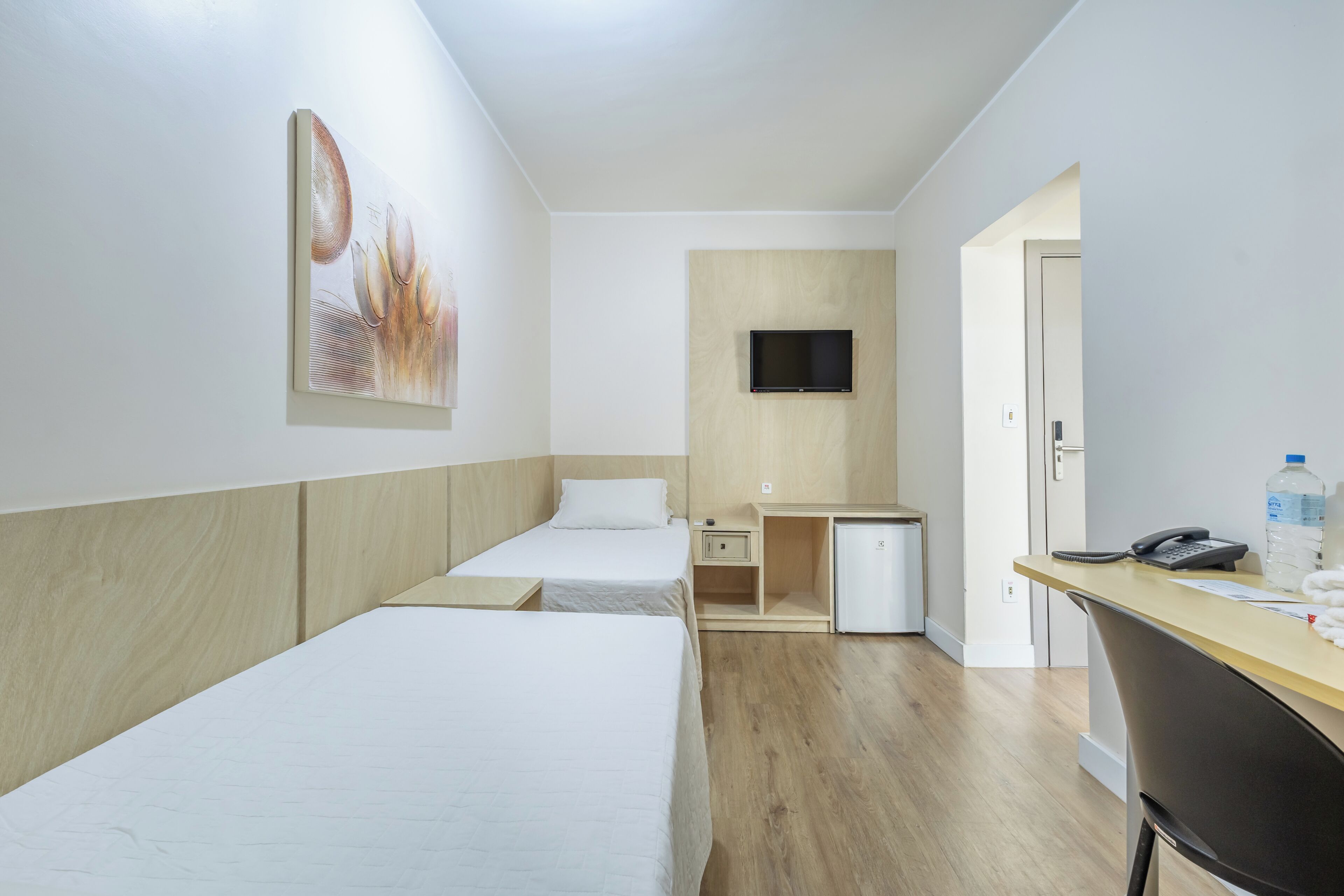 Habitación de hotel con dos camas individuales, un escritorio con televisor, un refrigerador pequeño y una puerta que da a otra habitación.