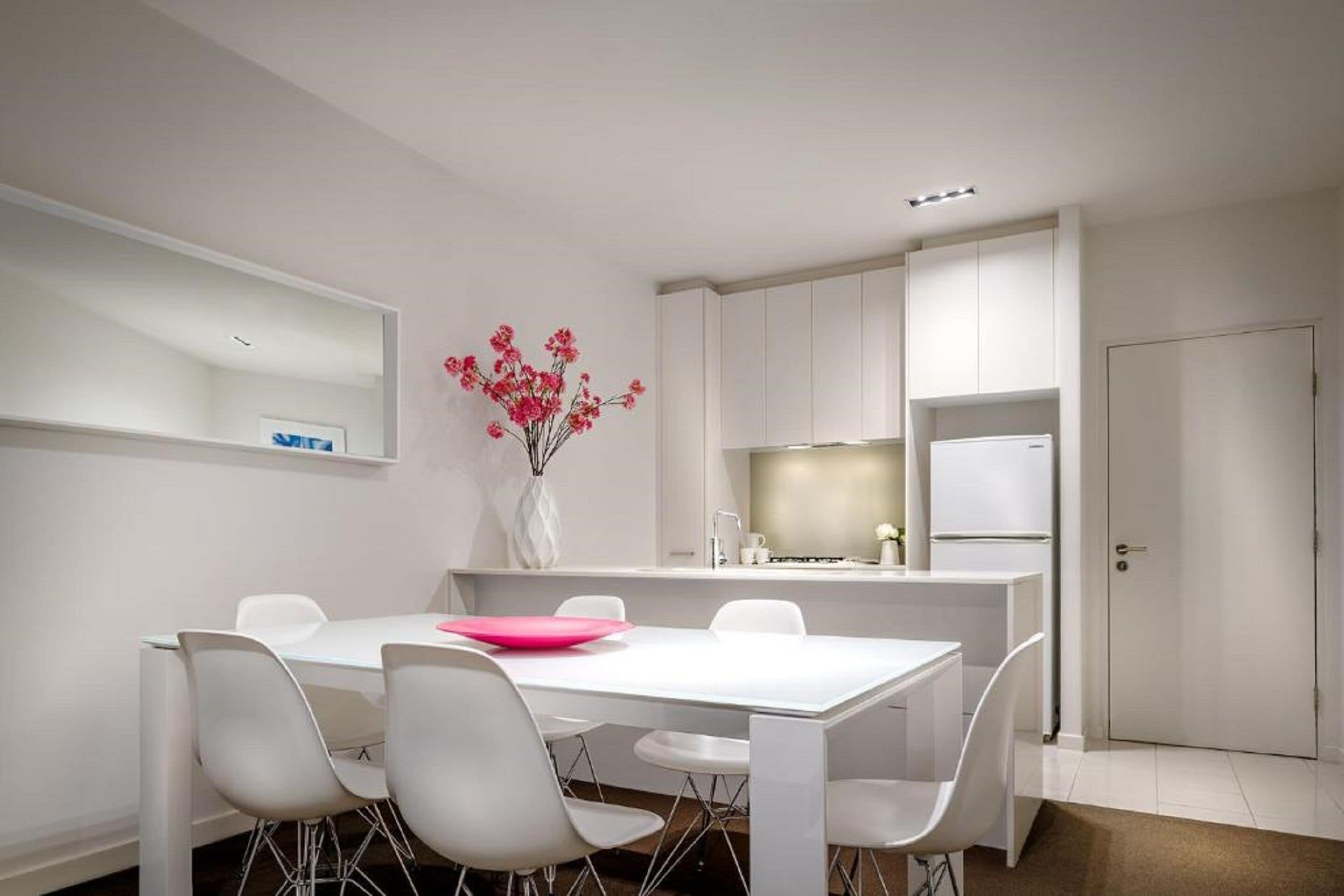 A modern kitchen with white cabinetry, a dining area with a white table and chairs, and a vase with pink flowers on the wall.