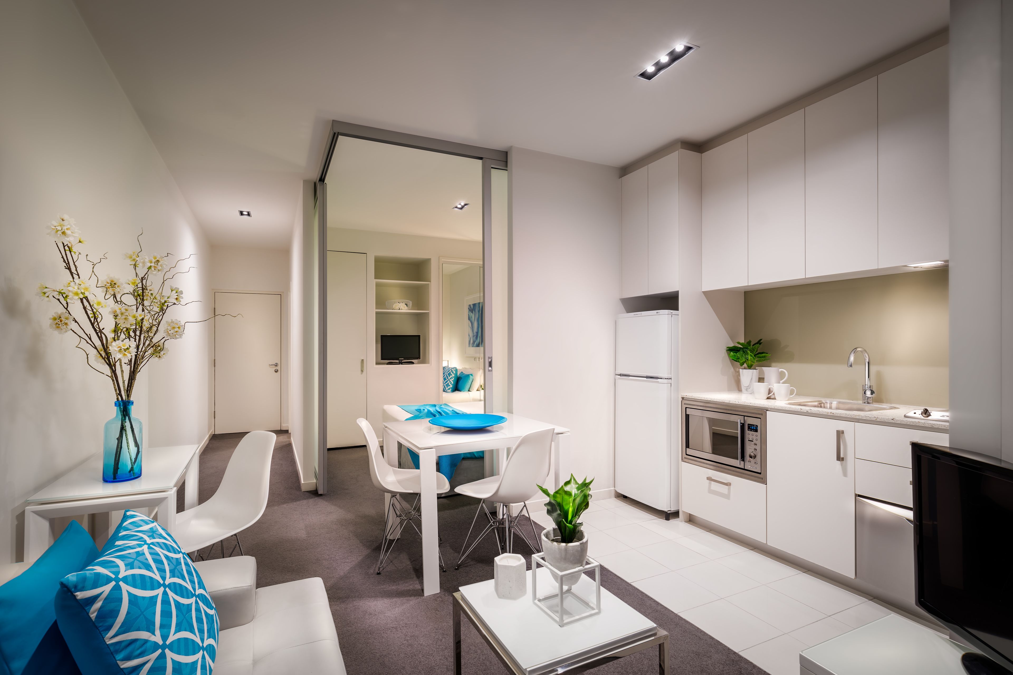 Una cocina moderna con armarios blancos, horno empotrado y microondas. Hay una zona de comedor con una mesa y sillas blancas, y una mesita de centro con una planta.