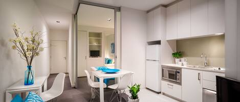 A modern kitchen with white cabinets, a built-in oven, and a microwave. There is a dining area with a white table and chairs, and a small coffee table with a plant.