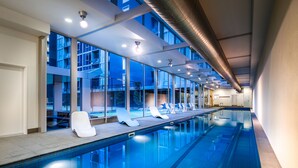 A long indoor swimming pool with clear blue water, white lounge chairs, and large windows offering views of the outside. - Milano Serviced Apartments (Melbourne)