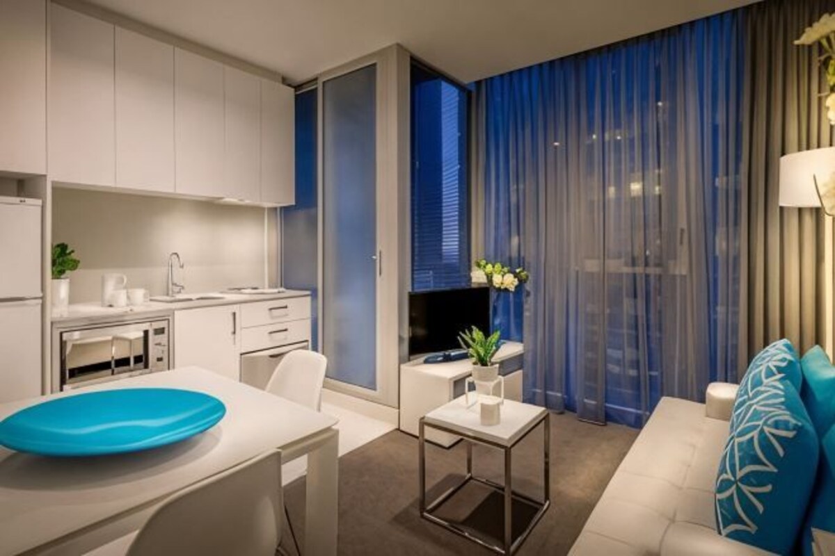 Une chambre d’hôtel moderne équipée d’une kitchenette, d’un coin repas, d’un canapé et d’une grande fenêtre avec des rideaux bleus.