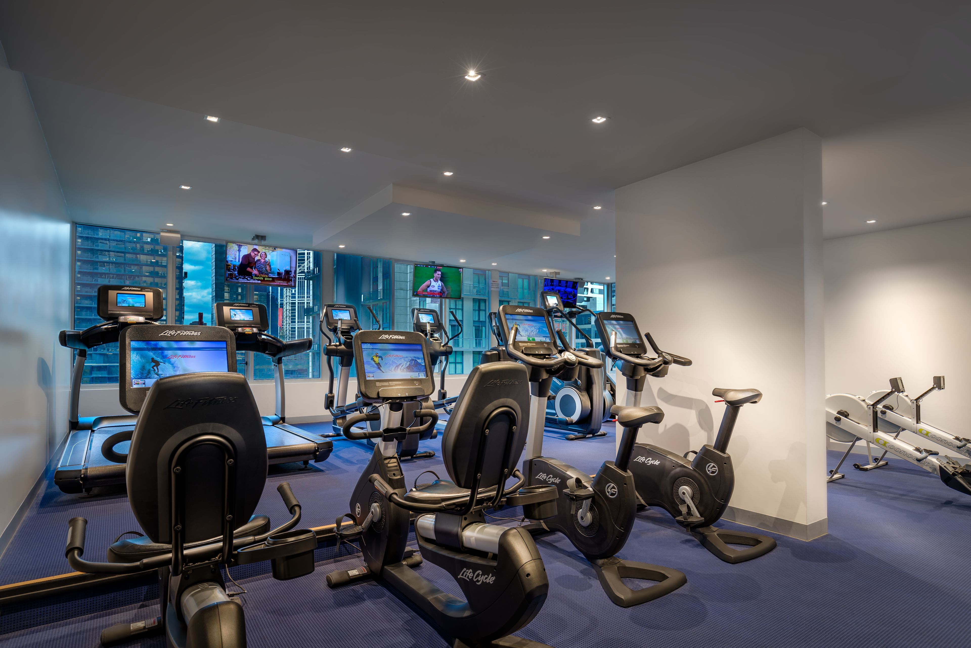 A gym with multiple exercise machines, including treadmills and stationary bikes, and large windows with city views.