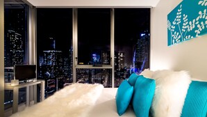 A modern bedroom with a city view, a white fluffy area rug, and teal pillows on a white sofa. - Milano Serviced Apartments (Melbourne)