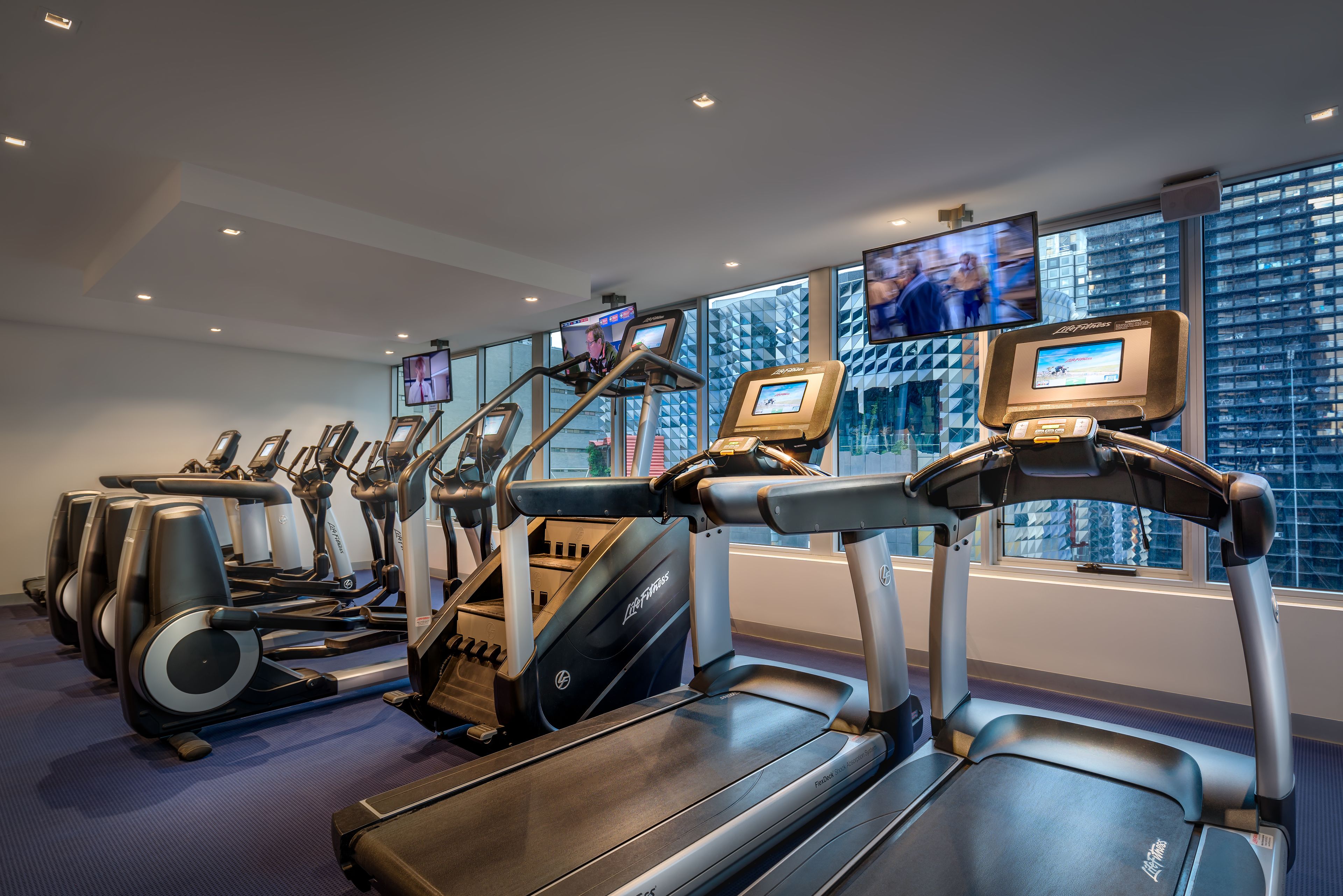 Une salle de sport équipée de tapis de course, d’elliptiques et de vélos d’appartement, dotée d’écrans numériques et de grandes fenêtres offrant une vue sur la ville.