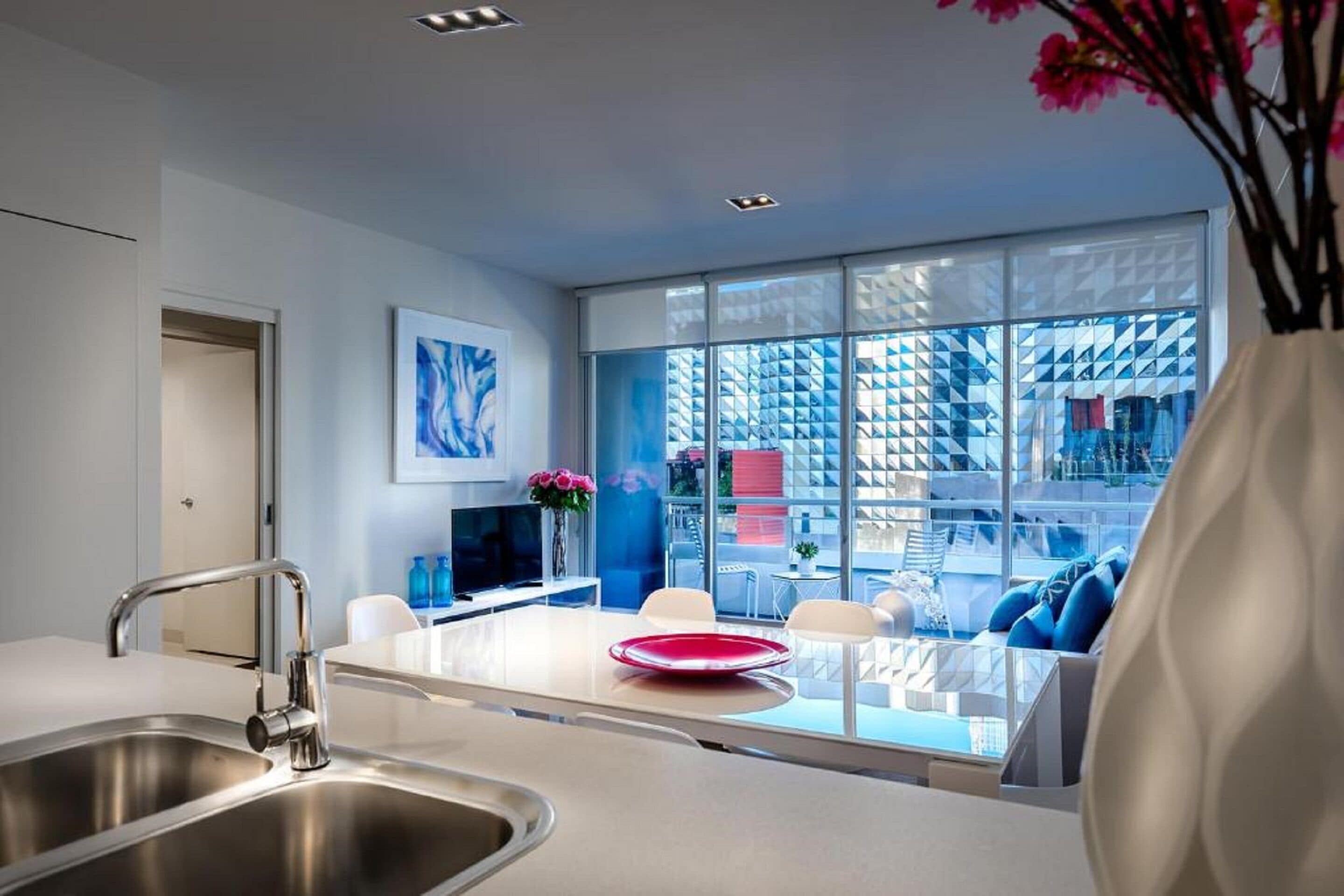 A modern kitchen with a stainless steel sink, a dining area with a glass table and white chairs, and a large window with a geometric view.