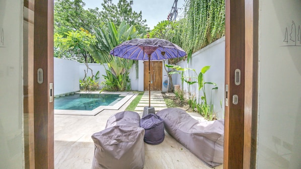 A pool area with a wooden door, a small pool, and a pathway leading to a door. There are two bean bags and a purple umbrella.