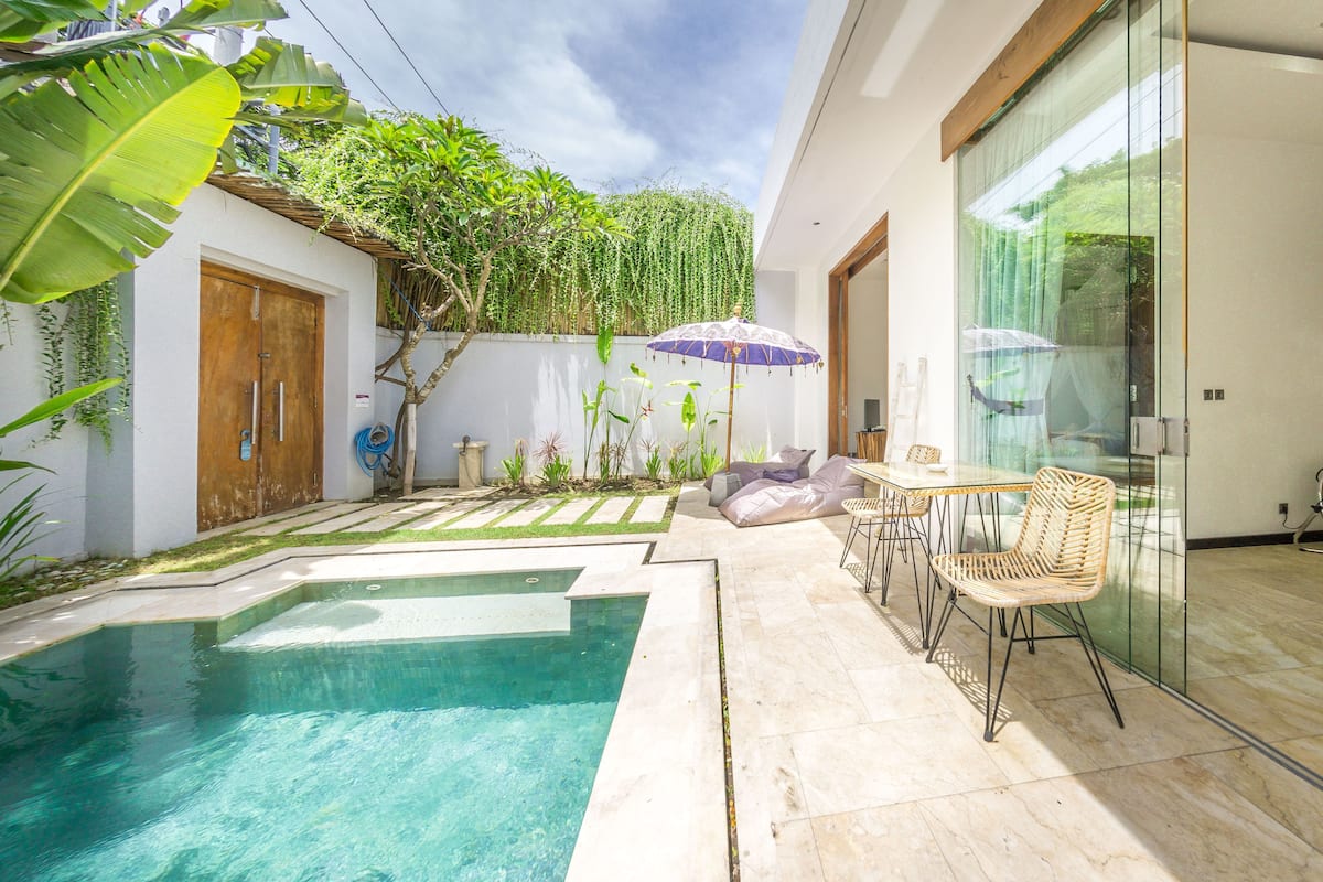 A modern outdoor pool area with a glass wall, a seating area with cushions, and a small kitchen area.