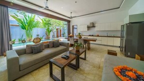 Una sala moderna con sofá, mesa de centro y comedor con banco. Se observa una cocina con armarios blancos y un refrigerador.