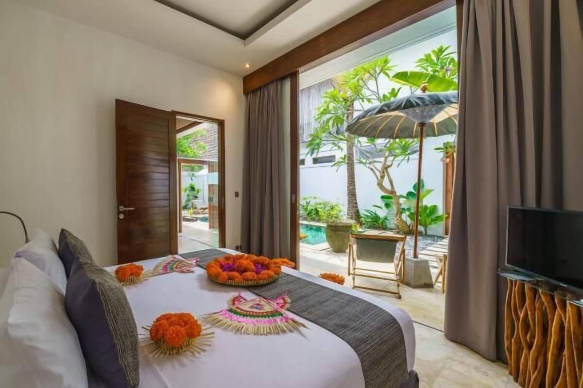 A hotel room with a bed, a TV, a balcony with plants, and a door leading outside.