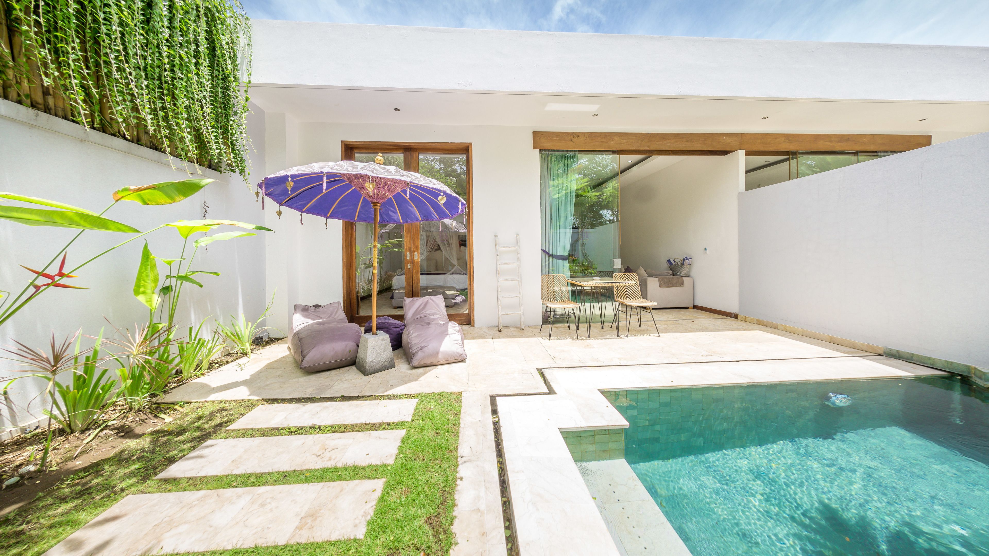 A modern outdoor pool area with a glass door leading to a dining area, surrounded by greenery and a clear blue pool.