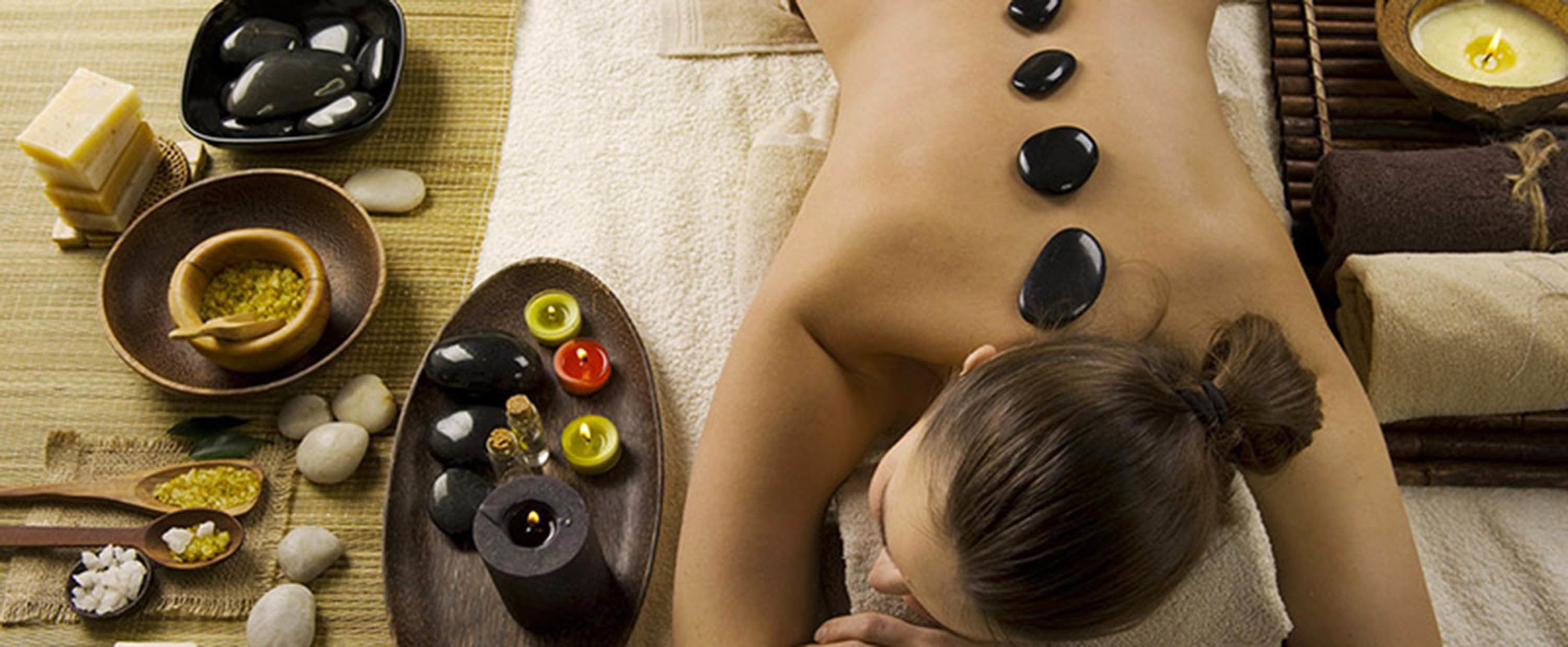 Une personne reçoit un massage aux pierres chaudes, avec des pierres, des bougies et des huiles essentielles, sur un tapis.