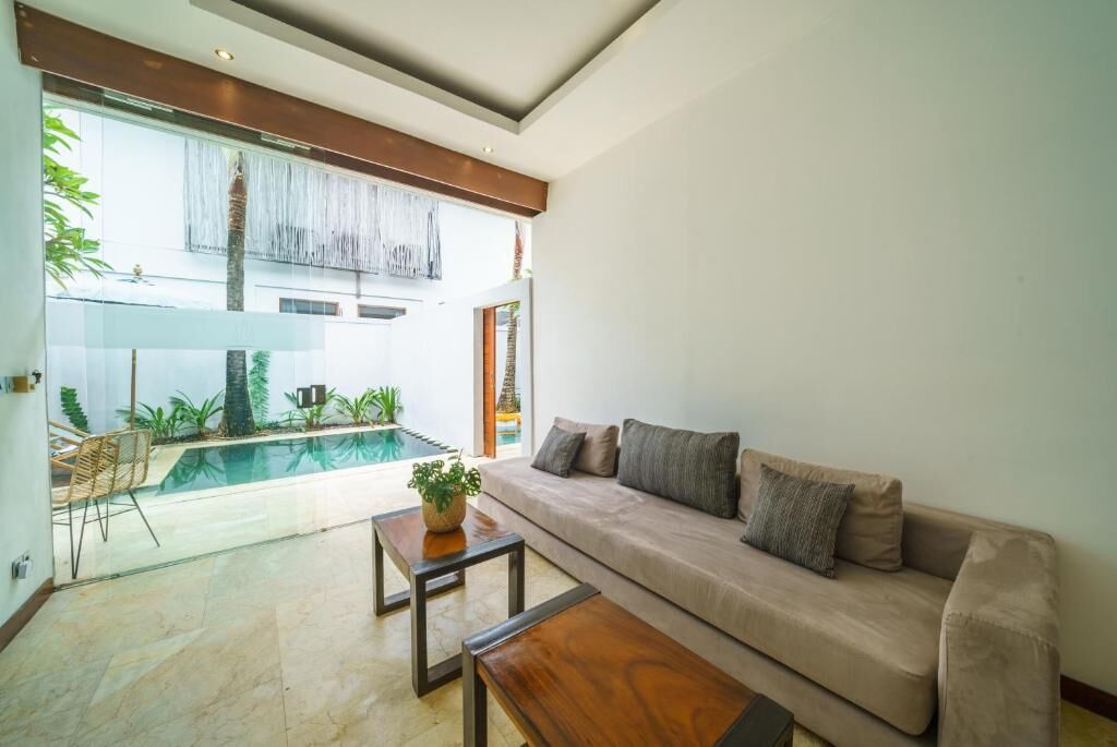 Una sala de estar moderna con un sofá color beige, una mesa de centro de madera y una mesa de comedor de vidrio. Se puede ver una piscina a través del muro de vidrio, y la habitación tiene un diseño minimalista con luz natural.
