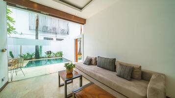 Una sala de estar moderna con un sofá color beige, una mesa de centro de madera y una mesa de comedor de vidrio. Se puede ver una piscina a través del muro de vidrio, y la habitación tiene un diseño minimalista con luz natural.