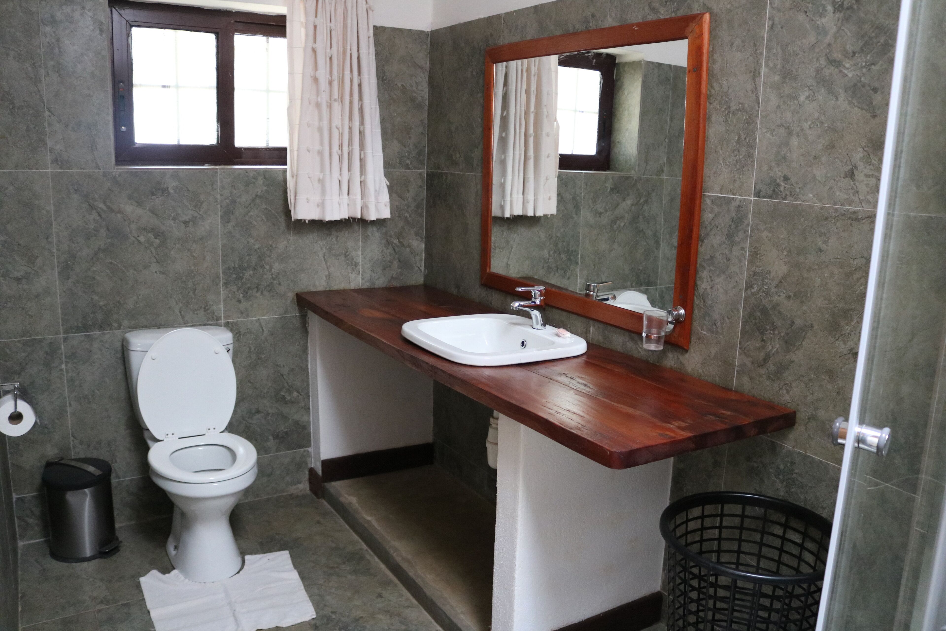 Un baño con inodoro blanco, tocador de madera con lavamanos blanco, espejo y cubo de basura negro.