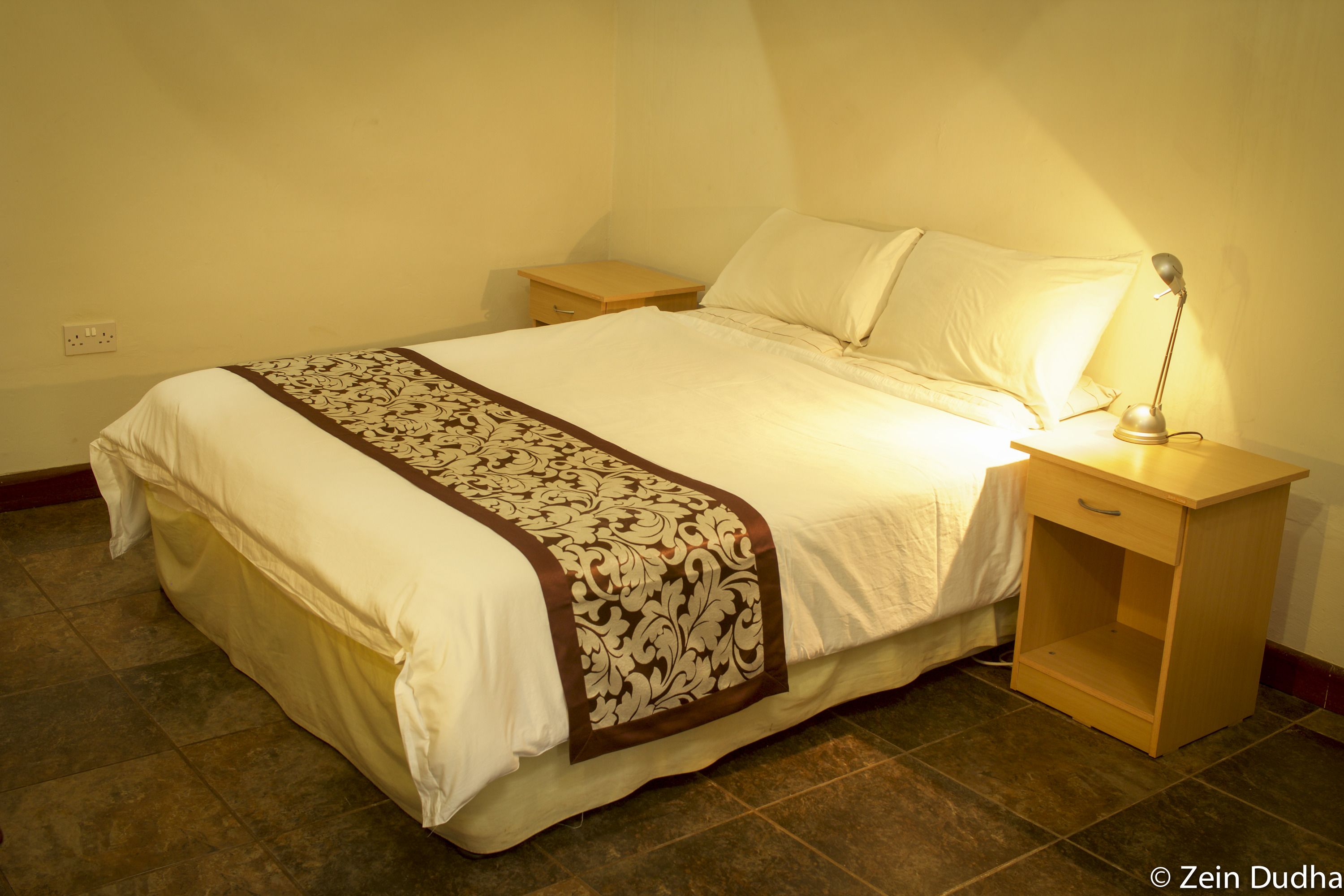Un lit bien fait, recouvert d’une couvre-lit à motifs, avec une lampe de chevet et une table de nuit en bois.