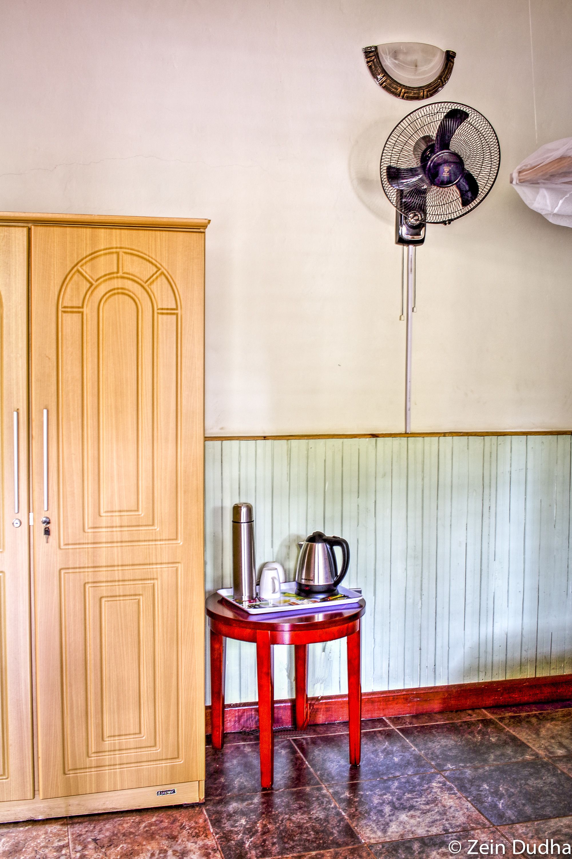 A wooden wardrobe, a red table with a kettle and a fan, and a tiled floor.