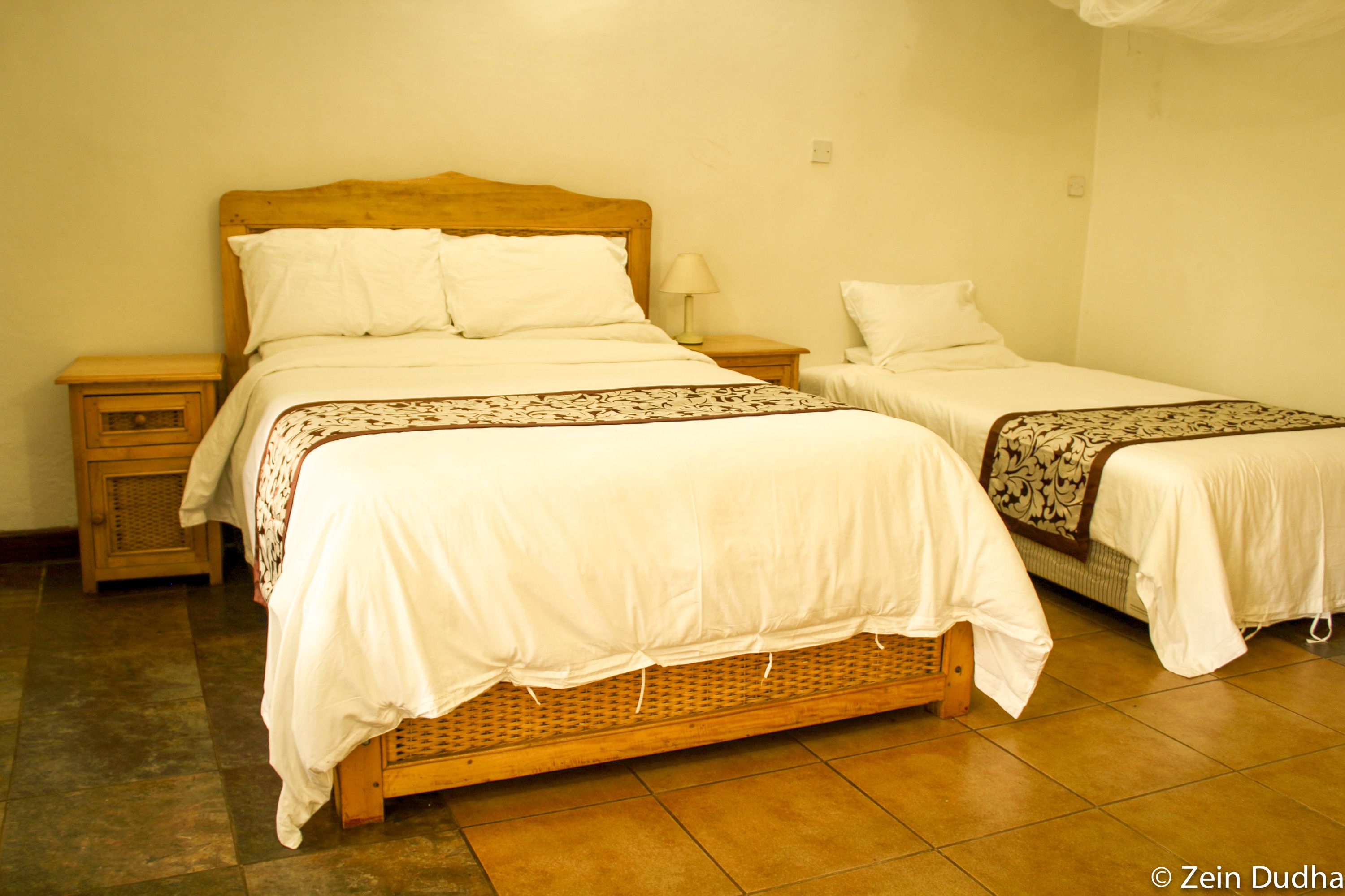 Una cama doble con cabecera de madera y una cama individual con estructura de madera en una habitación con suelo de baldosas.