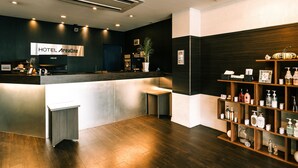Un espace d’accueil d’hôtel au design moderne, comprenant un comptoir d’accueil, un écran numérique et un panneau mural indiquant « ZONE HÔTEL ». On y trouve des étagères en bois avec des articles de toilette et une plante en pot.