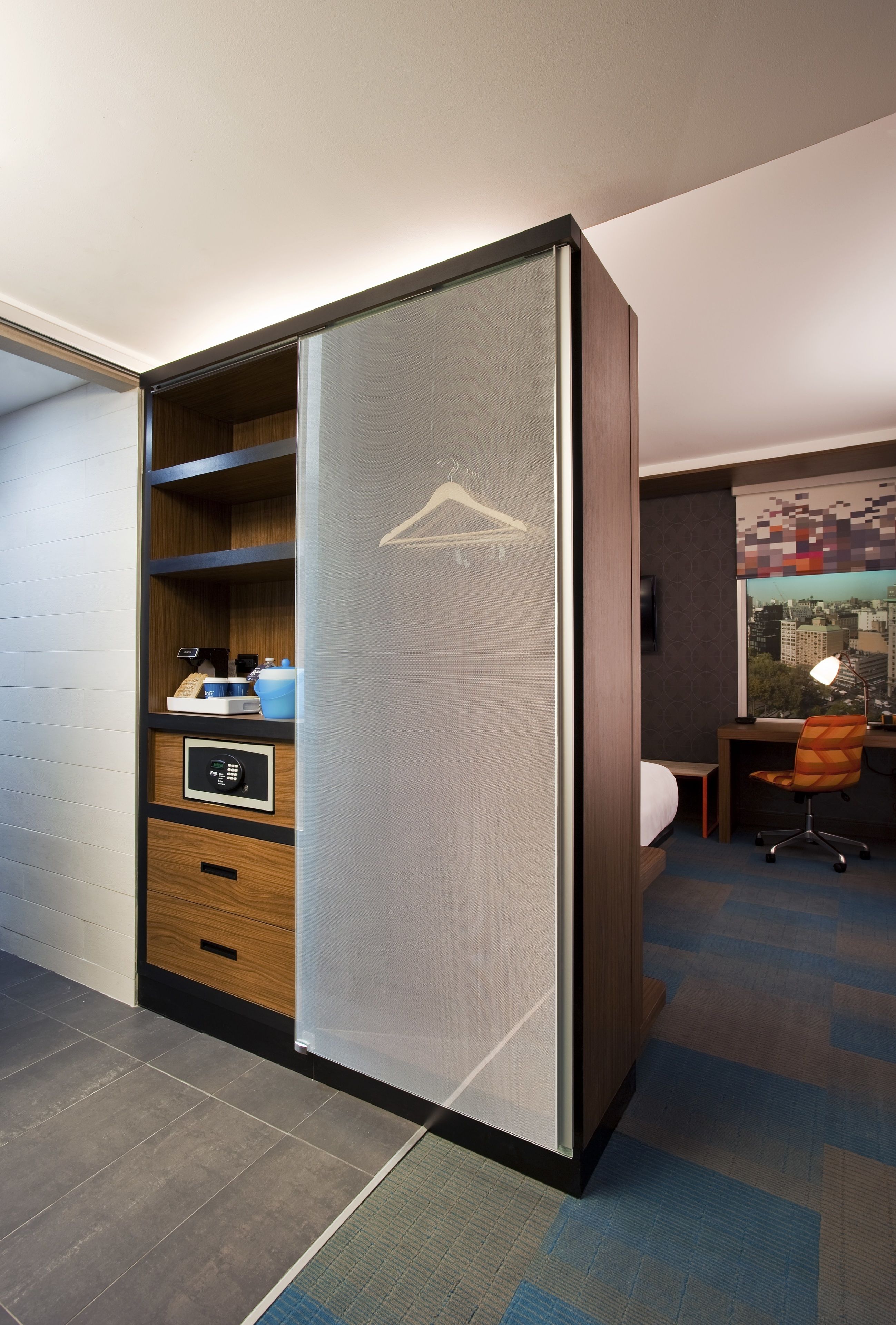 A hotel room with a sliding wardrobe, a desk with a chair, a bed, and a cityscape view through the window.