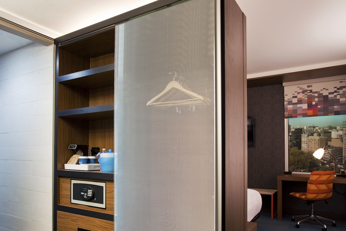 Une chambre d’hôtel équipée d’une armoire à portes coulissantes, d’un bureau avec une chaise, d’un lit et offrant une vue sur le paysage urbain par la fenêtre.