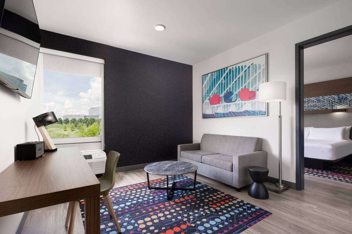 A modern hotel room with a sofa, a small round table, a desk with a lamp, a patterned rug, and a large window with a view of greenery.