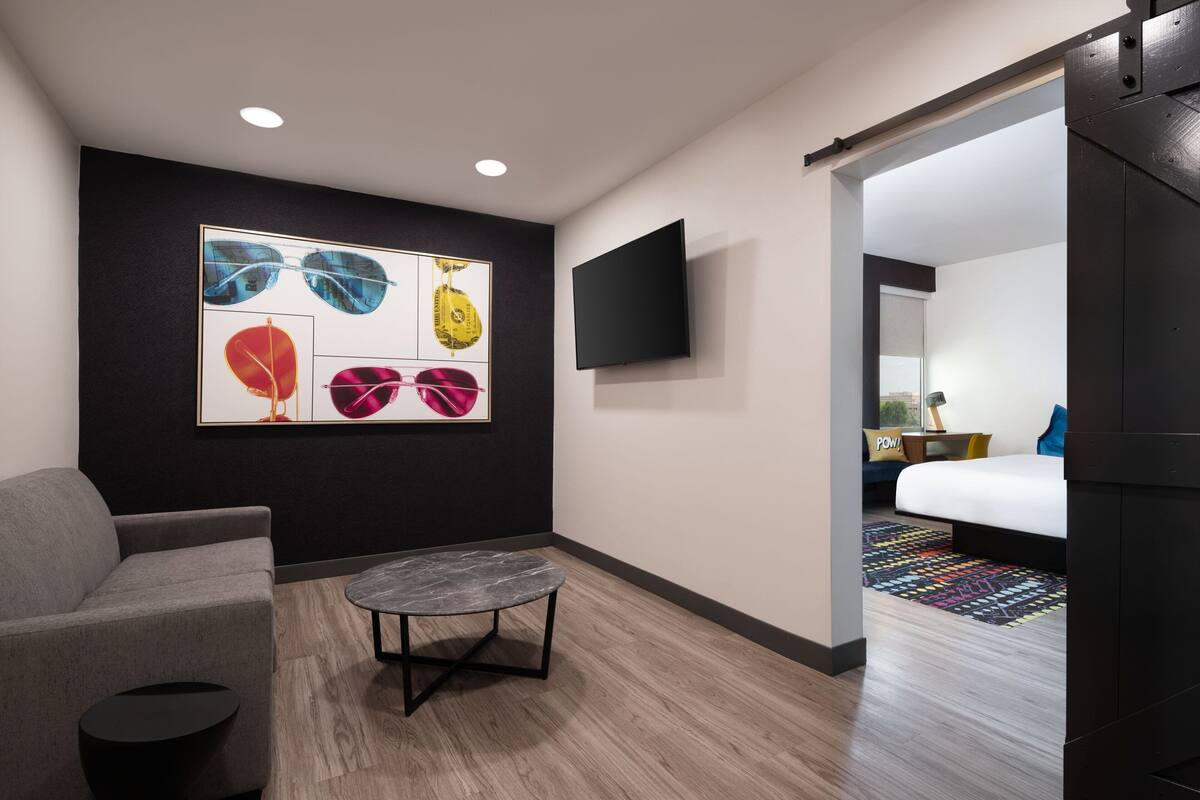 A modern hotel room with a gray sofa, a small round table, a large abstract painting on the wall, a flat-screen TV, and a bed with a colorful rug.