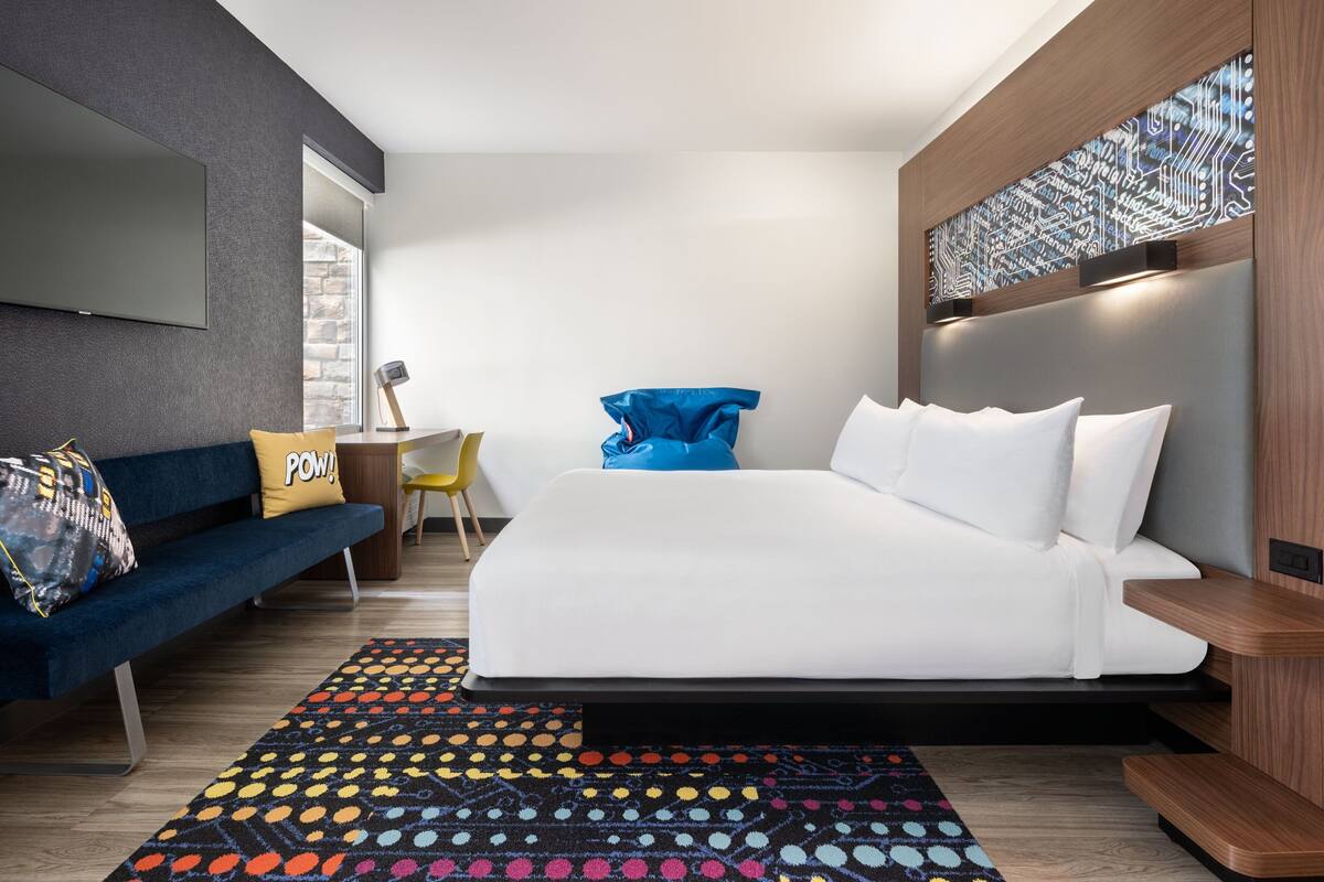 A modern hotel room with a large bed, a sofa, a desk, and a colorful rug.
