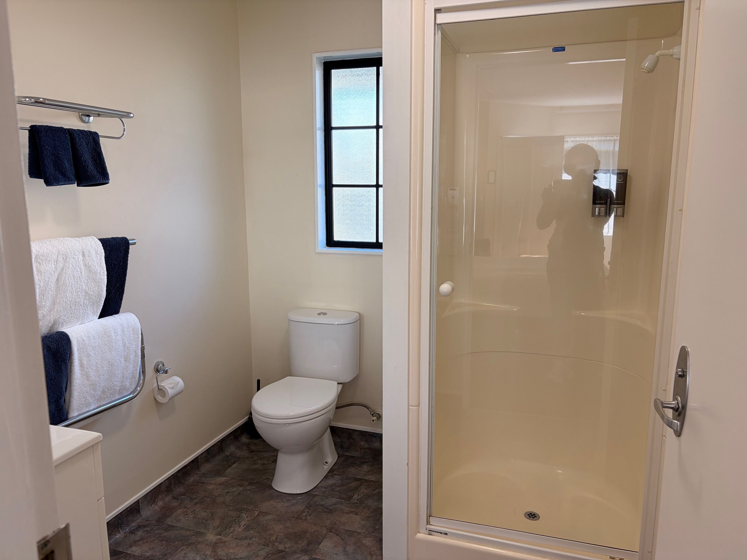 Une salle de bain comprenant des toilettes, un porte-serviettes avec des serviettes, une fenêtre et une douche fermée par une paroi de verre.