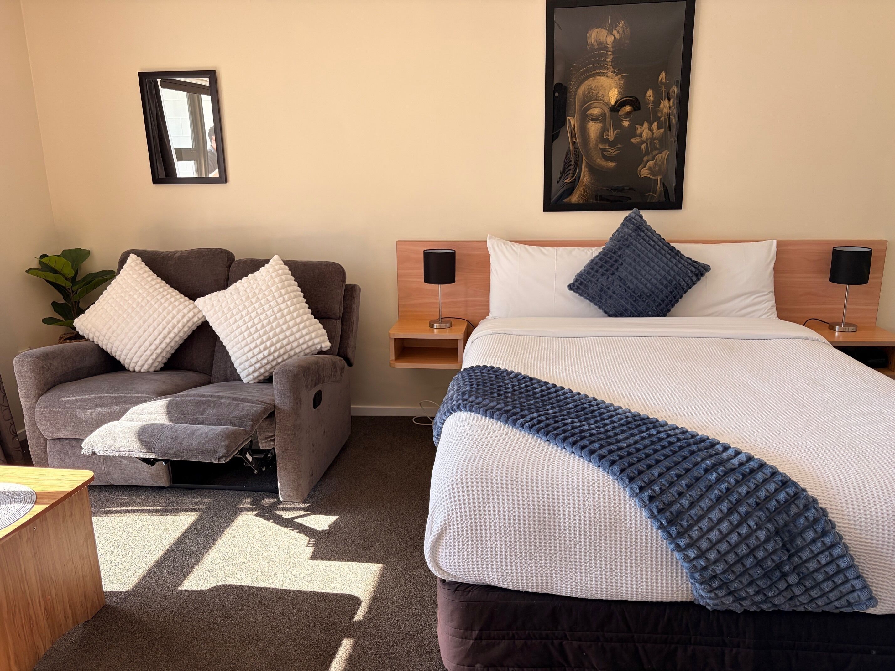 A hotel room with a bed, a chair, a nightstand, a lamp, a wall-mounted picture, and a plant.