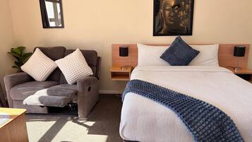 A hotel room with a bed, a chair, a nightstand, a lamp, a wall-mounted picture, and a plant.