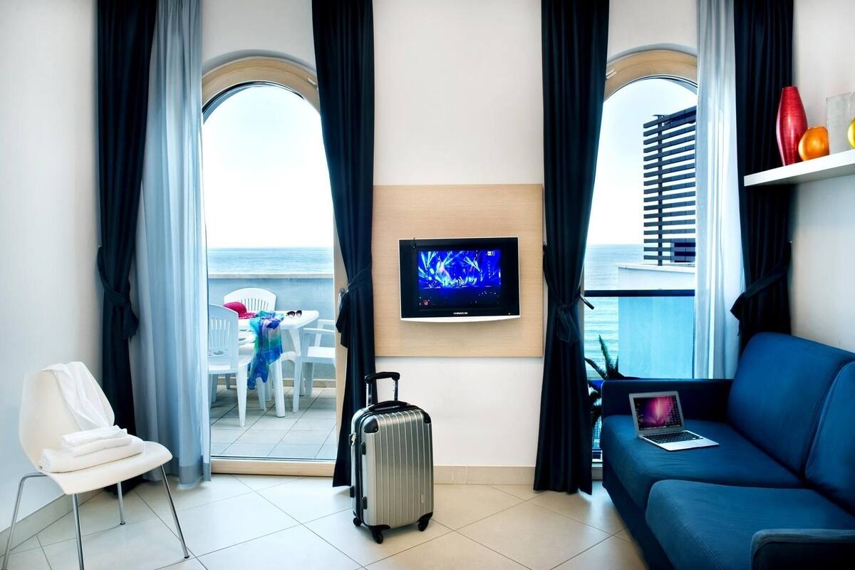Habitación de hotel con balcón, televisión, sofá, silla y maleta.