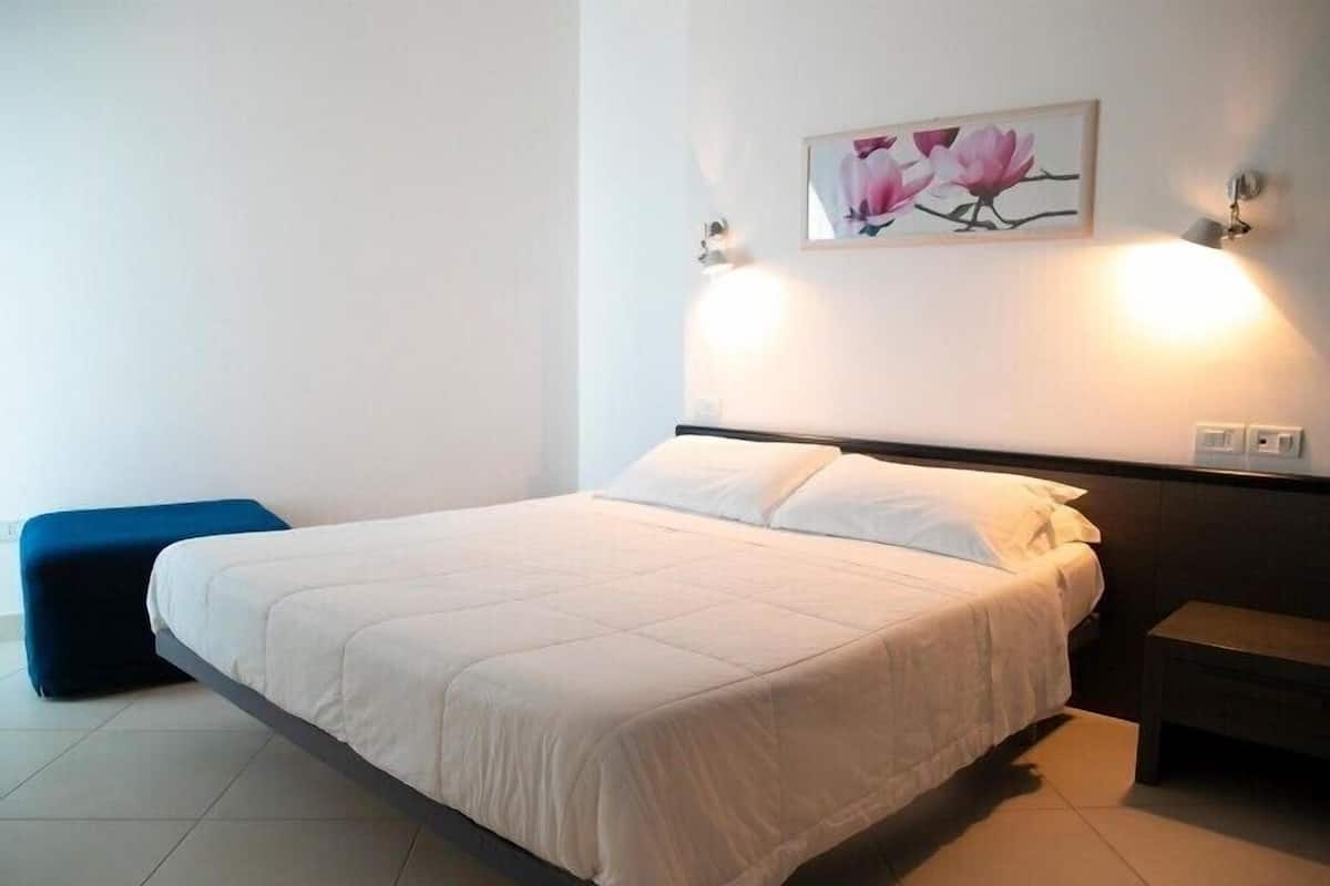 Una cama impecablemente tendida con ropa blanca, un banco azul y un cuadro con flores rosadas colgado en la pared.