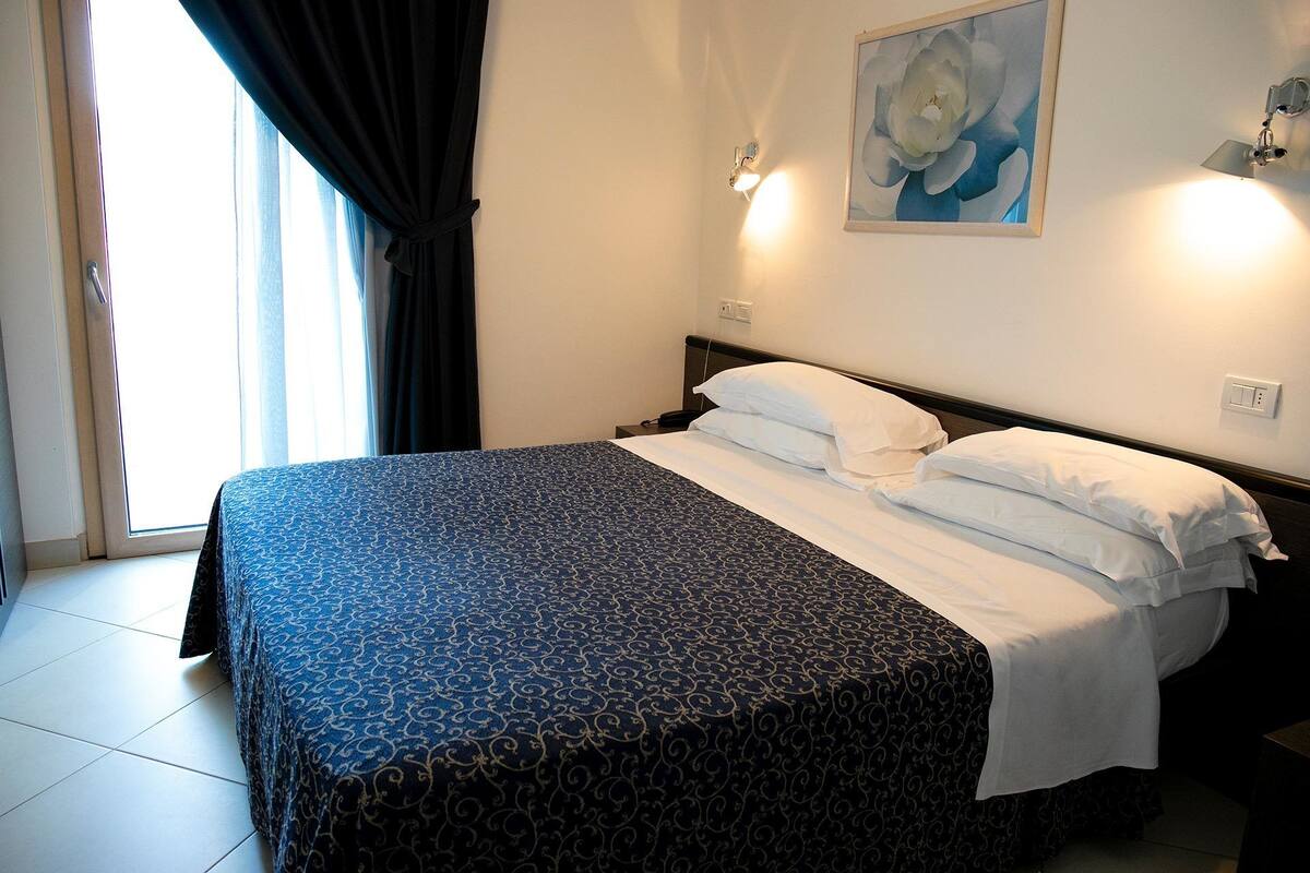 Una habitación de hotel con una cama, una mesita de noche, un cuadro y una ventana con cortinas.
