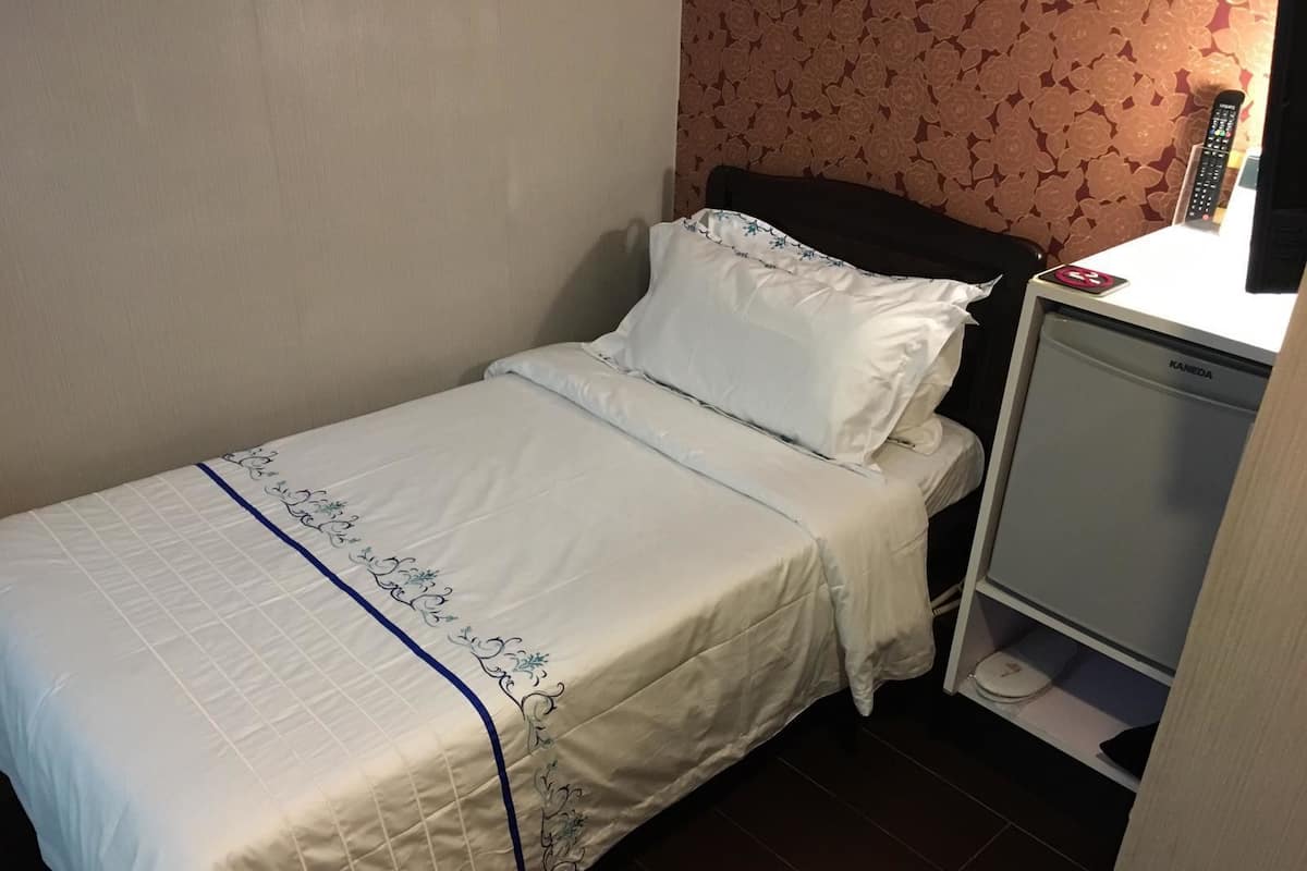 A single bed with a white bedspread and blue trim, a nightstand with a remote control, and a small cabinet with a scale.