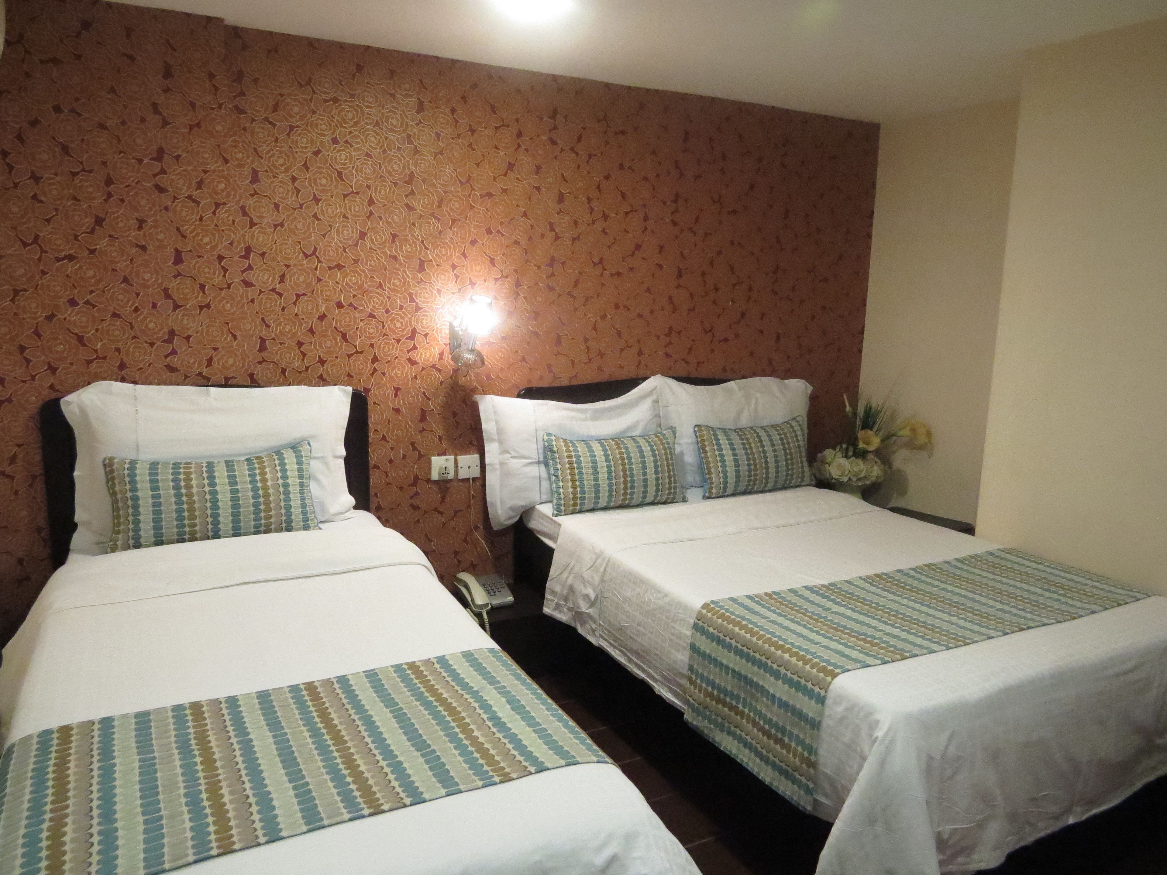 A hotel room with two beds, each with a patterned bedspread and pillows. There is a wall-mounted telephone and a vase with flowers on a nightstand.