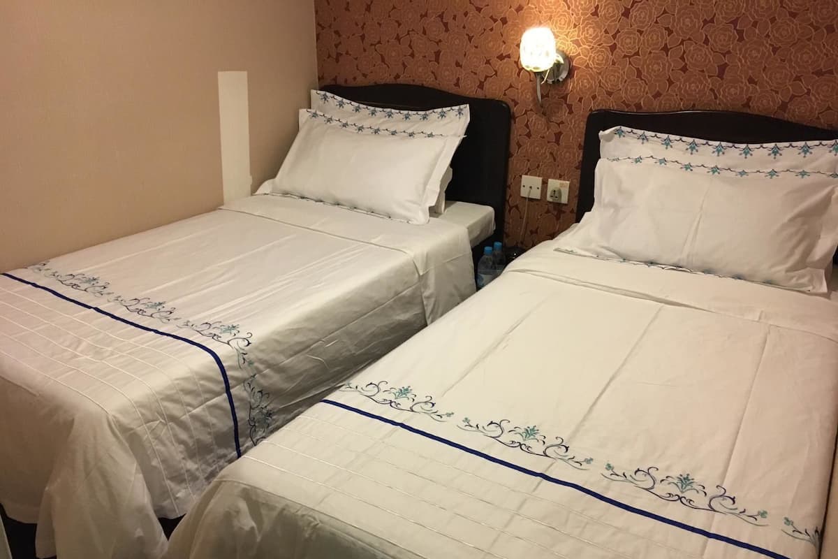 Two single beds with white linens and blue floral embroidery, a wall-mounted light, and a floral-patterned wallpaper.