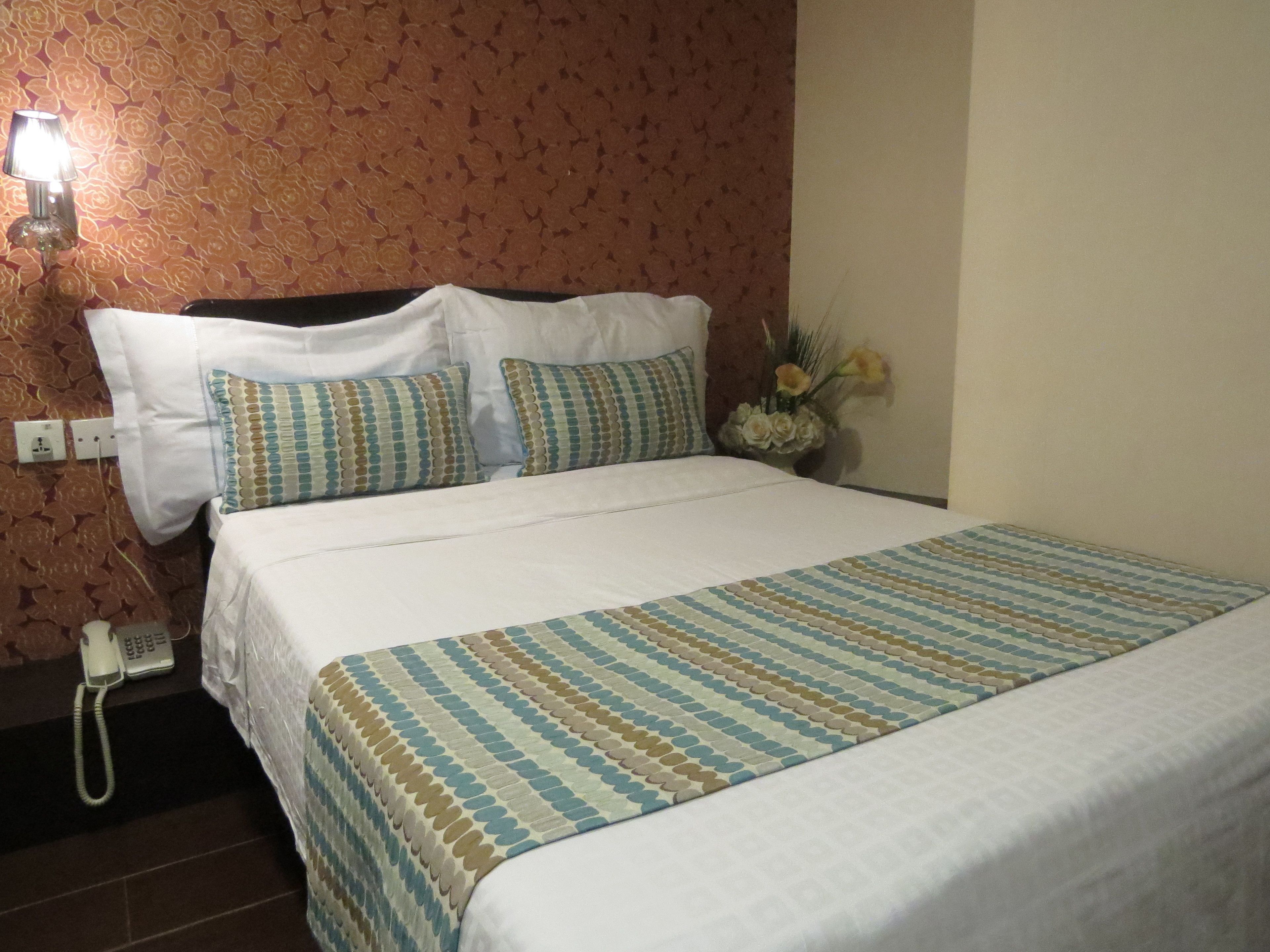 Habitación de hotel con cama, ropa de cama blanca, almohadas estampadas, teléfono y arreglo floral.