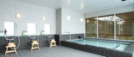 A modern bathroom with a large bathtub, three wooden stools, and a window with a view of a garden.