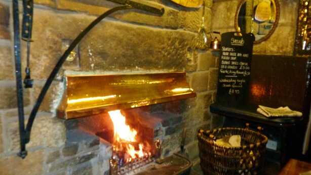 Una chimenea de piedra con fuego visible, un menú en pizarra que enumera alimentos y una cesta de mimbre en el suelo.
