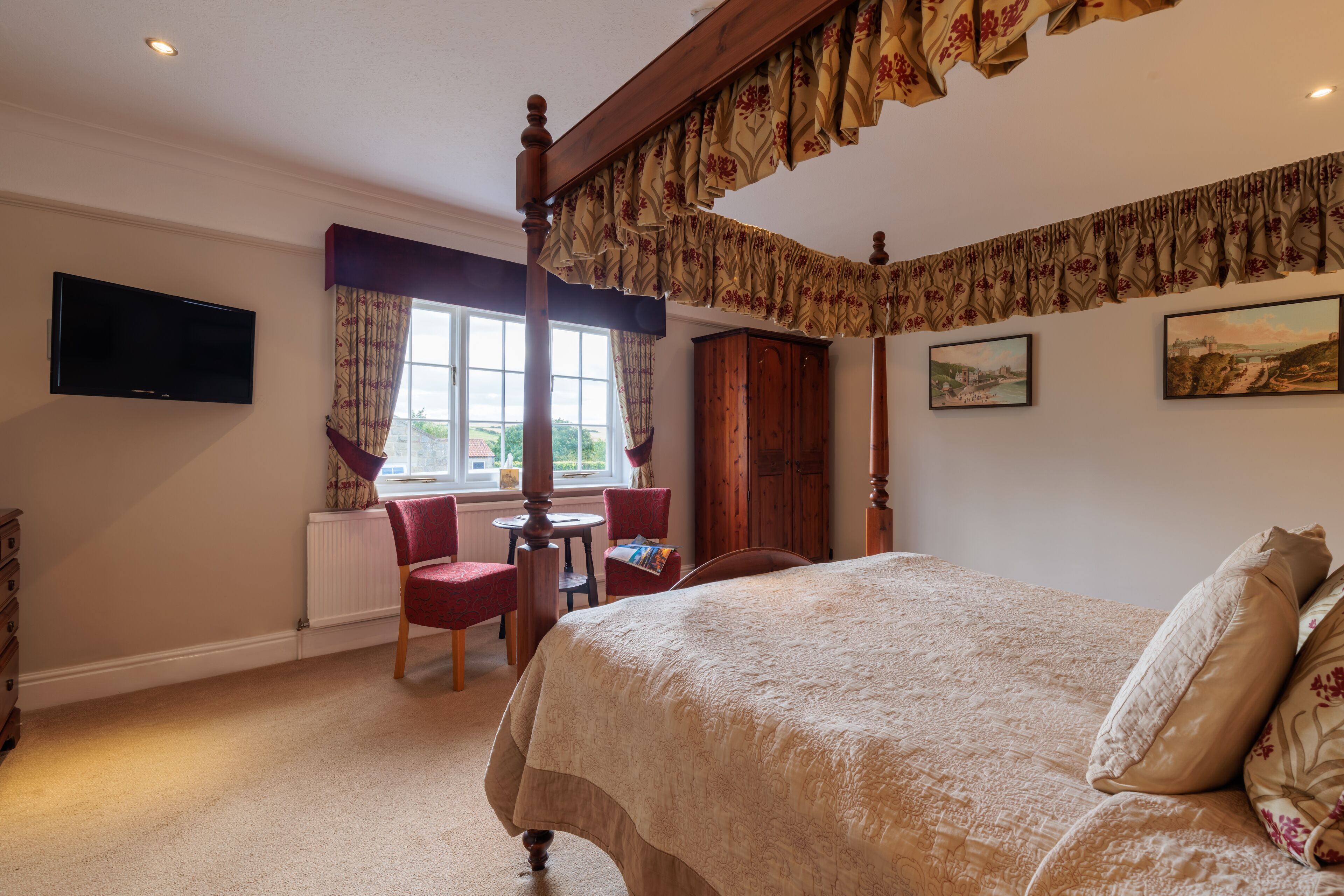 Een slaapkamer met een hemelbed, een aan de muur bevestigde televisie, een raam met gordijnen en uitzicht op het omliggende landschap.