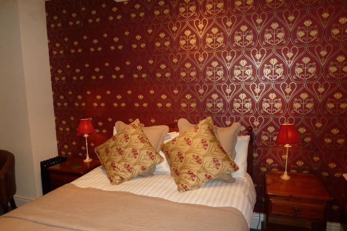 Quarto de hotel com papel de parede estampado, uma cama com duas almofadas e duas mesas de cabeceira com luminárias.