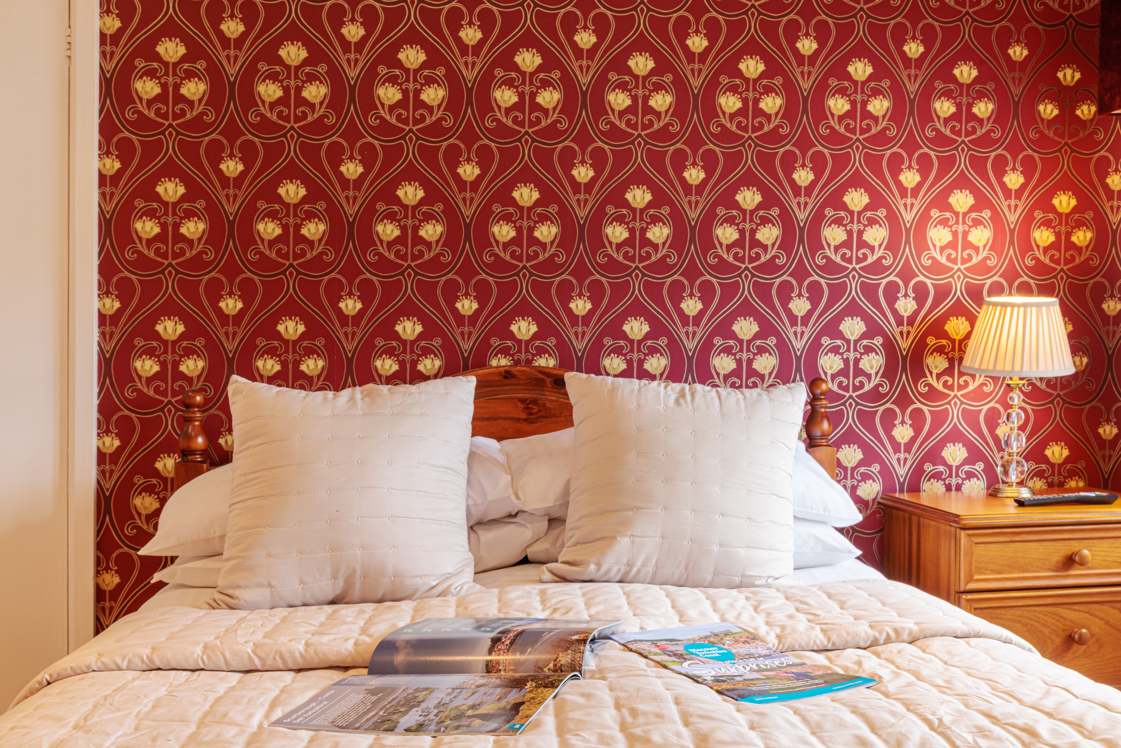 Cama bem arrumada com roupa de cama e travesseiros brancos, cabeceira de madeira e mesa de cabeceira com abajur e um livro.