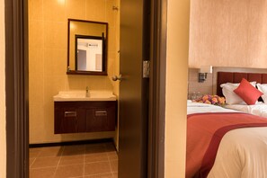 A hotel room with a bathroom featuring a sink and mirror, a bed with a red and white bedspread, and a nightstand with a lamp and flowers.