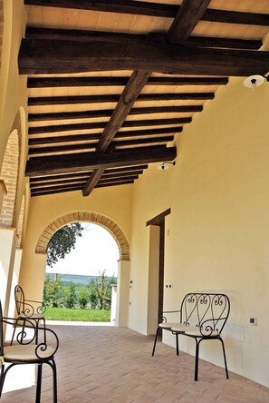 Un patio al aire libre con techo de madera y vista a la vegetación. Se observan dos sillas de metal y una puerta que da a otra habitación.