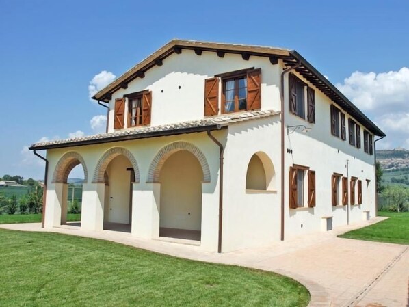 A two-story house with a white facade, brown shutters, and a tiled roof. It features a covered porch with arches and a paved driveway.