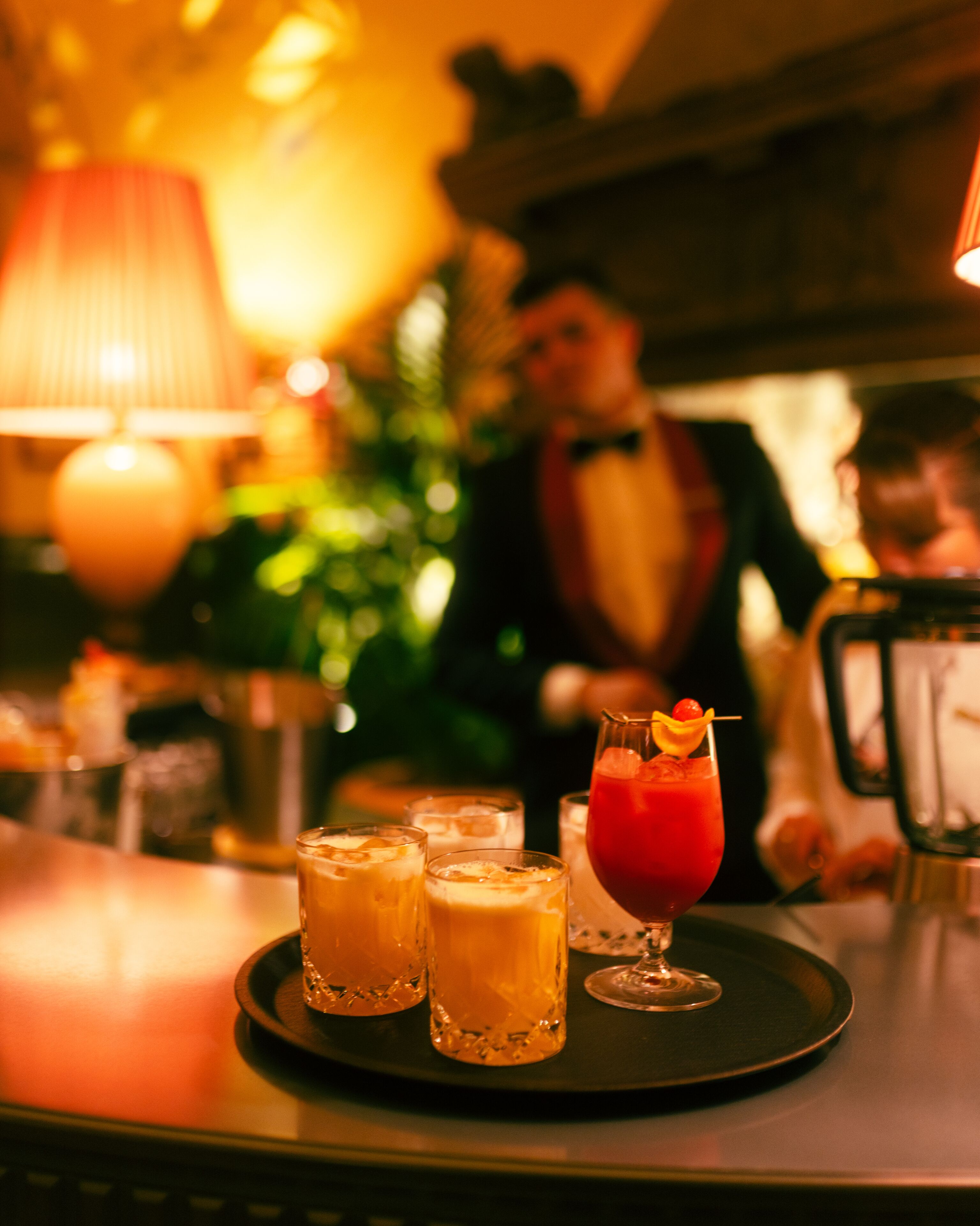 Une scène de bar avec un menu de boissons, un verre à cocktail garni d’une cerise et un barman en arrière-plan.