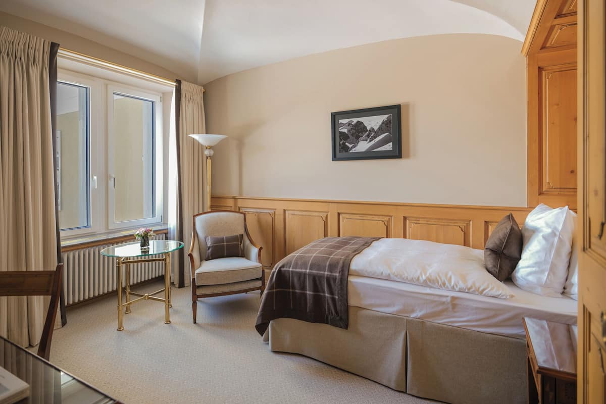 A hotel room with a bed, a chair, a small table, and a window with curtains.